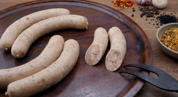 Vier feine Bratwürste angerichtet auf einem Schneidbrett und eine halbierte Bratwurst, bei welcher man die Würze im Teig sieht.